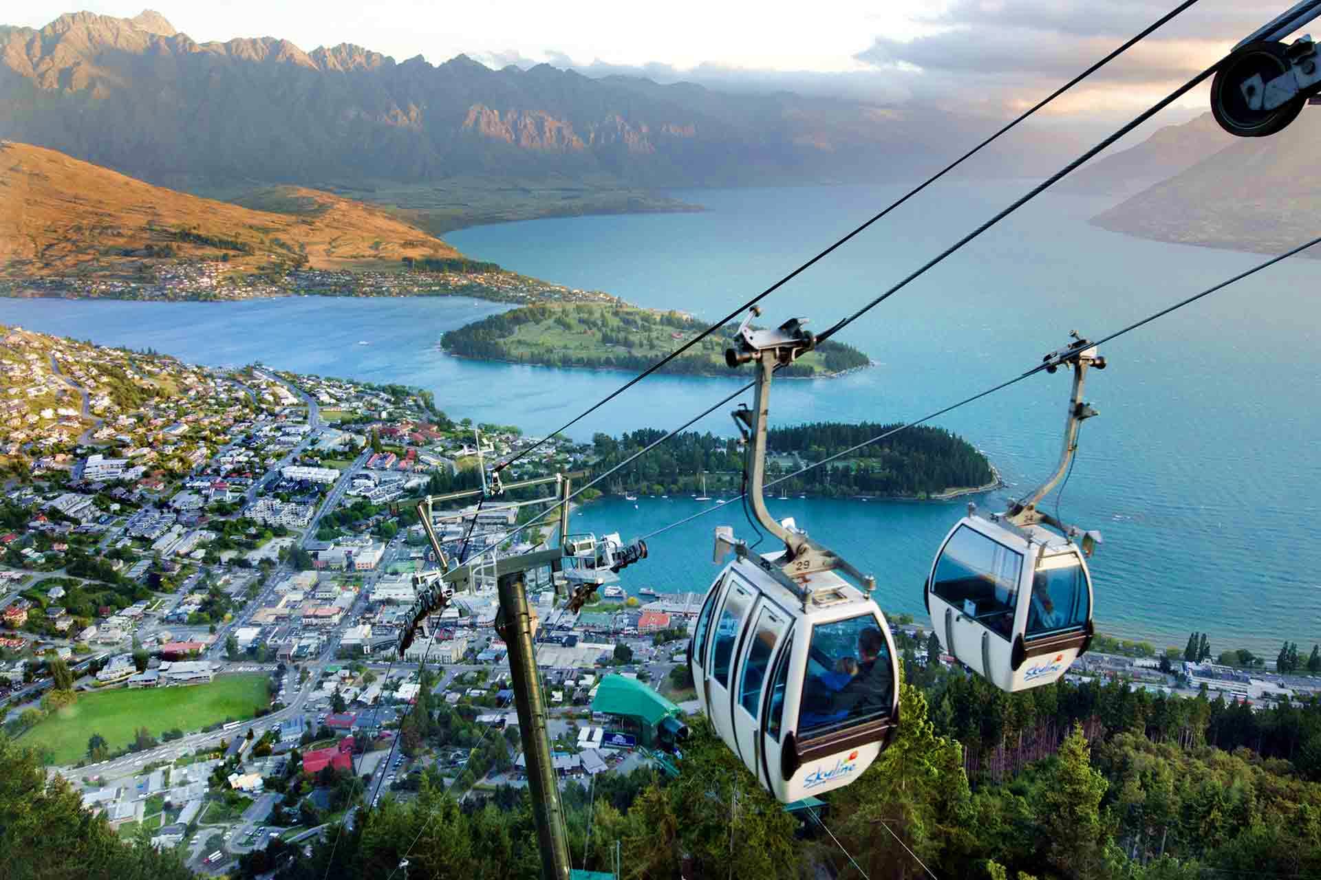 Gondola cable cars suspended above Lake Wakatipu with Queenstown city, mountains, and forested peninsulas below in New Zealan